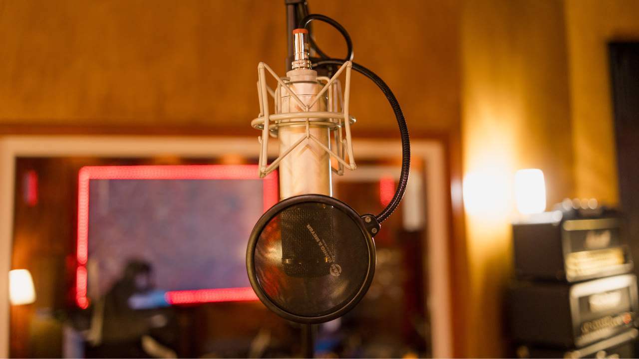 A close-up of a professional recording microphone with a pop filter in a studio, with a soundproof room in the background.