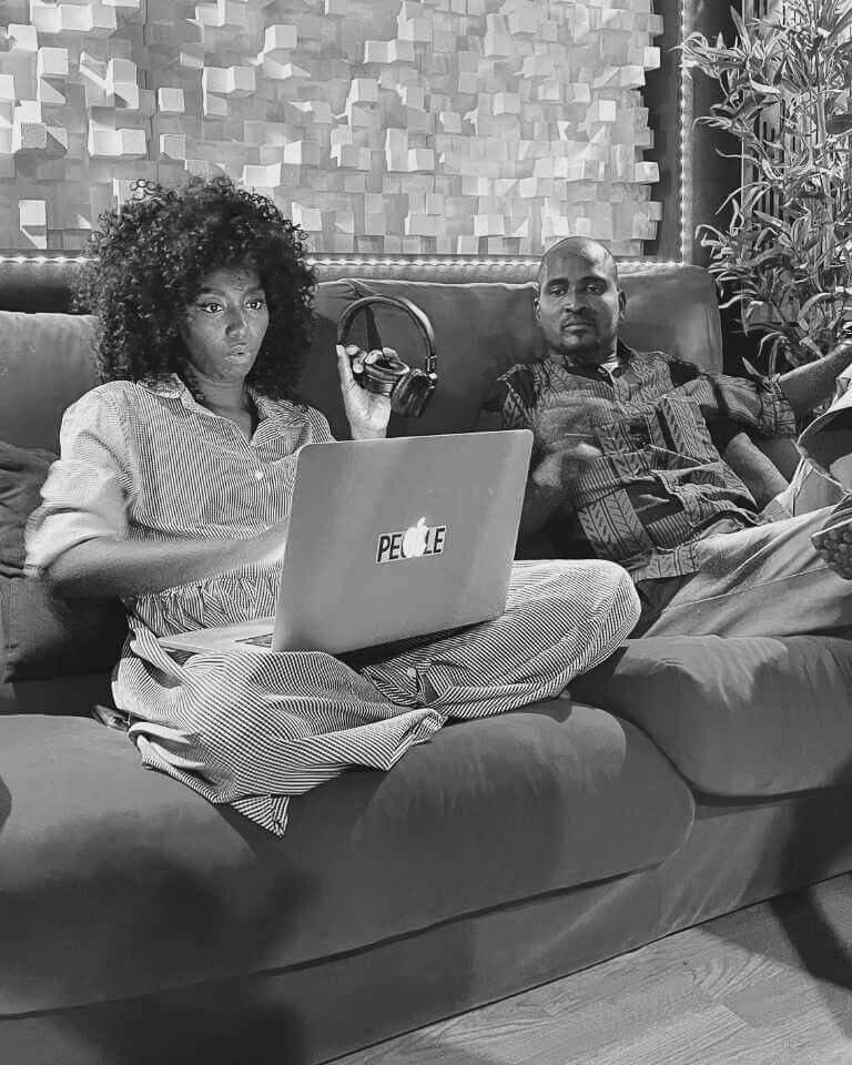 Two artists collaborating on a red couch in a music studio, one working on a laptop and the other holding headphones, with acoustic panels in the background.