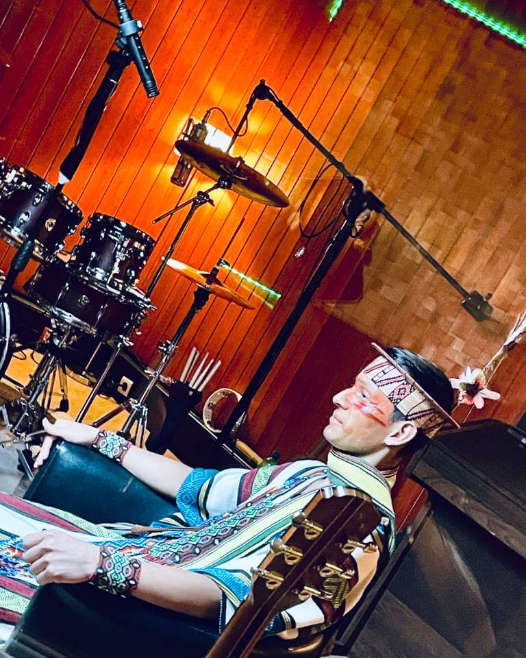 An artist in traditional attire during a recording session, seated in a professional studio with a drum kit, microphones, and wooden acoustic panels.