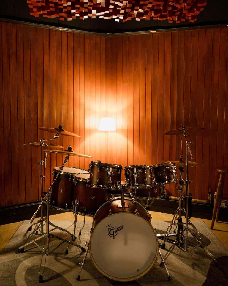 A professional drum set in a live room with wooden acoustic paneling and a diffused light lamp, creating a warm recording environment.