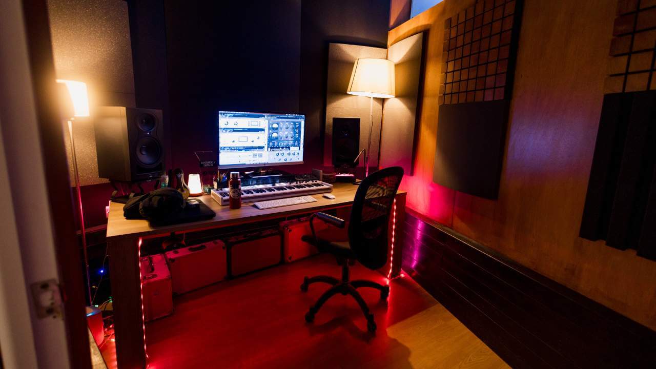 A modern music production workstation with a keyboard, monitor displaying plugins, and warm ambient lighting in a soundproof room.