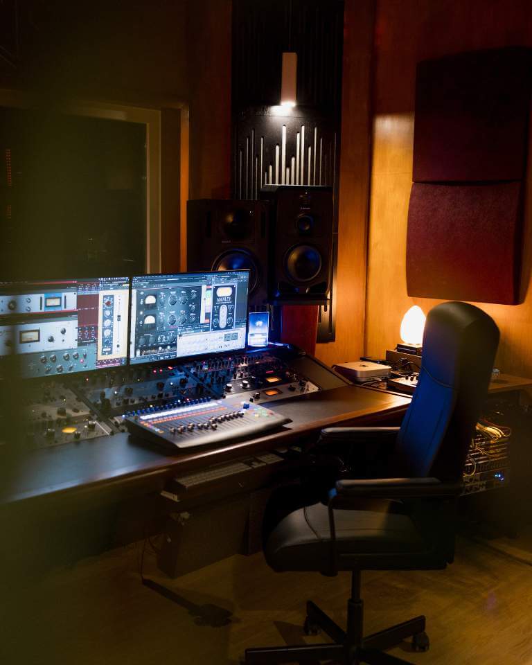 A professional mastering studio with a mixing desk, dual monitors displaying plugins, studio monitors, and a comfortable chair in a soundproof environment.