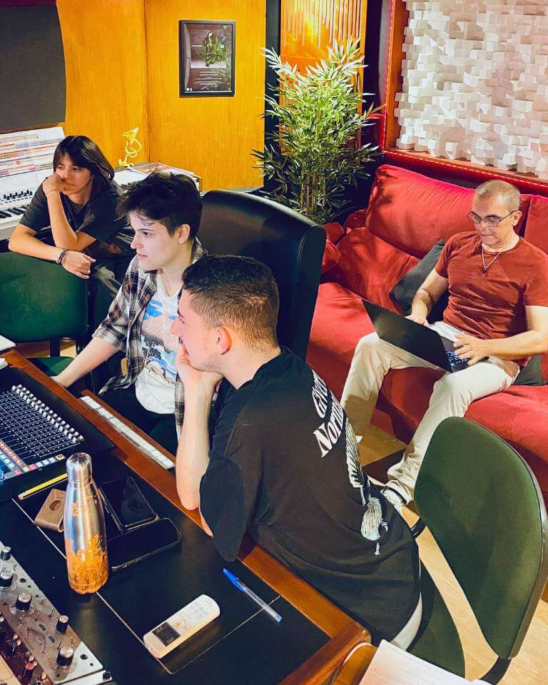 A collaborative music production and songwriting session in a studio, with producers and a songwriter working together at a mixing desk.