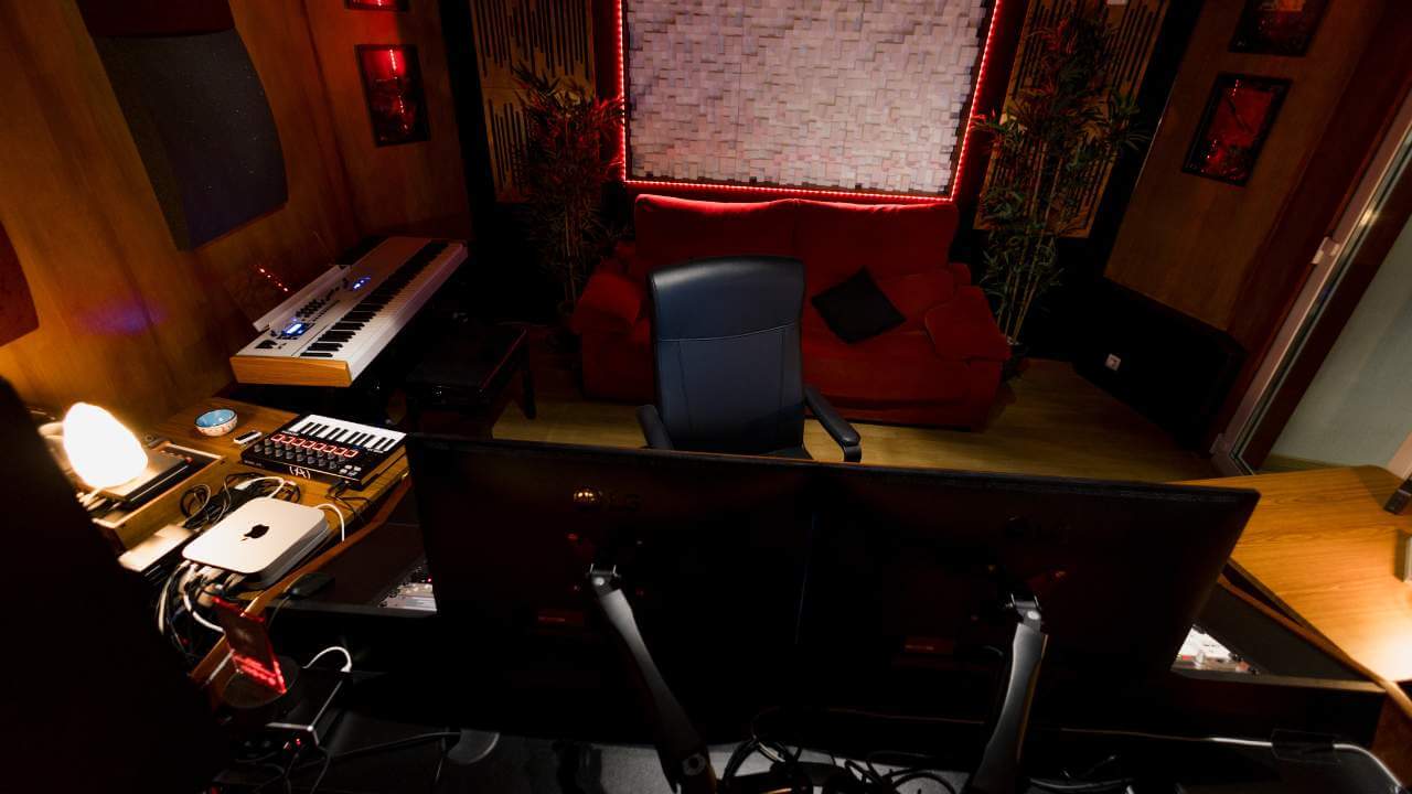 An overhead view of a professional music studio control room with dual monitors, keyboards, and a vibrant red couch in the background.