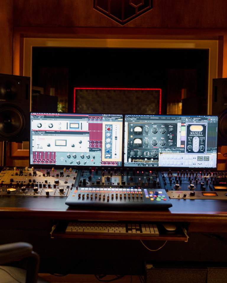 A professional mixing desk with dual monitors displaying audio plugins in a music studio, surrounded by studio monitors and ambient lighting.