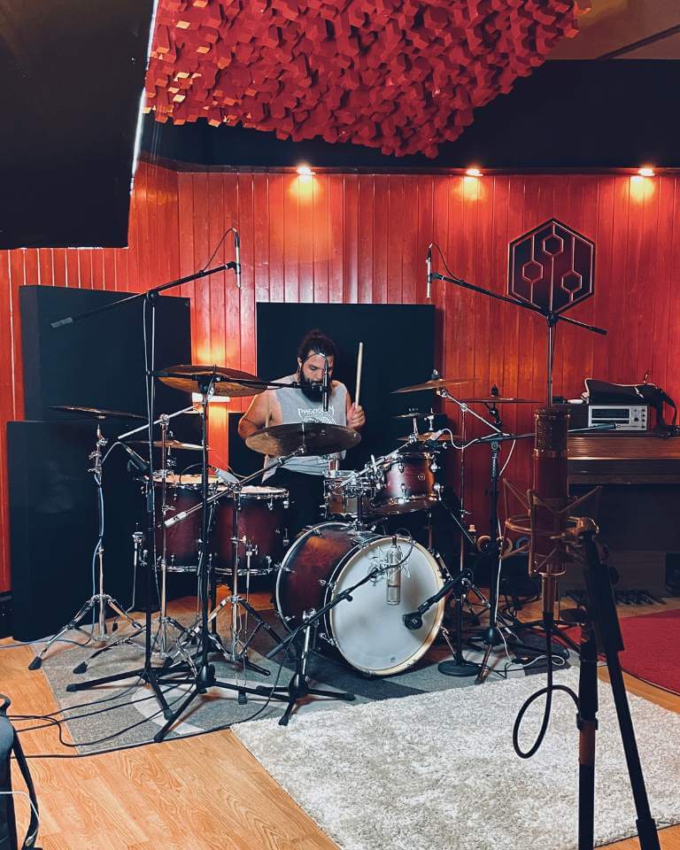 Drummer performing in a professionally treated recording studio with red acoustic panels and a fully mic'd drum kit.