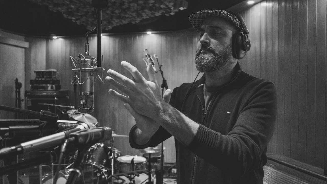 An artist wearing headphones during a vocal recording session in a professional studio, clapping in front of a microphone with wooden acoustic panels in the background.