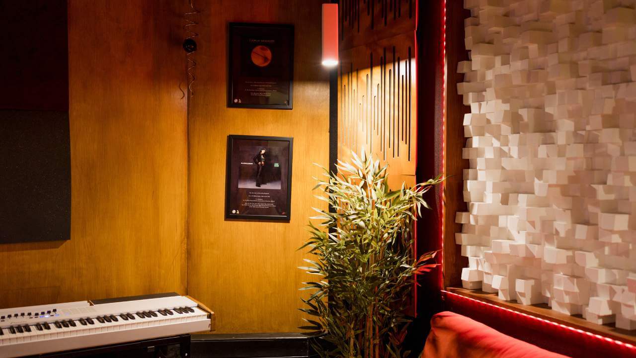 A stylish music studio gallery corner featuring a white keyboard, framed records on the wall, acoustic panels, and decorative lighting with vibrant red accents.